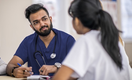 A multi-ethnic group of doctors are indoors in an office. They are gathered around a table for a meeting. One man is taking notes.