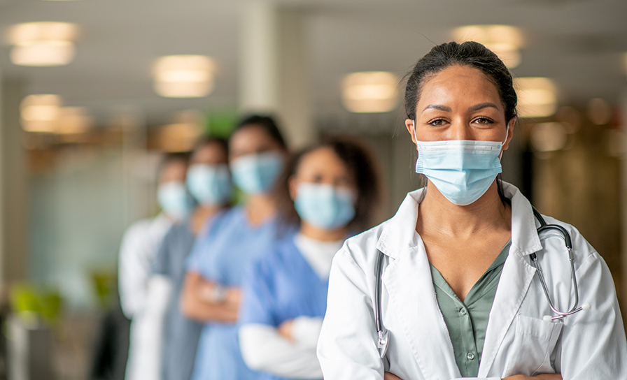 A diverse group of women stand in an institutional building in medical wear. A woman wearing scrubs stands at the forefront with a mask that is pulled down.