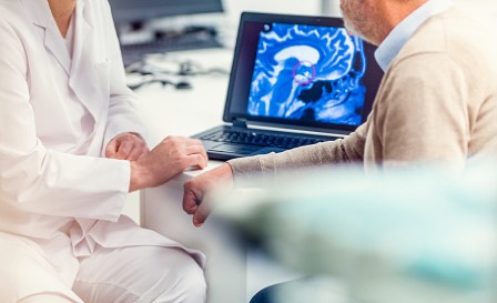 A doctor and his patient sitting at a table and looking at an MRI scan of the patientâ€™s brain which is showing a tumour.