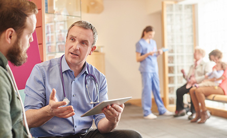 Male doctor holding a clipboard explaining something to a young adult male patient.